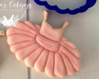 Op de afbeelding: Een roze ballet tutu koekje met gouden kraalaccenten op het lijfje. Een blauwe koekjesvorm is op de achtergrond. Het koekje is versierd met lichtroze glazuur en heeft een gedetailleerd ontwerp.