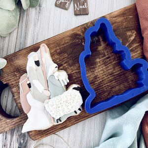 May include: A blue plastic cookie cutter shaped like a manger with a white and brown gingerbread cookie decorated with a nativity scene. The cookie features a shepherd, Mary, Joseph, and a lamb.