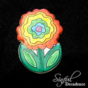 May include: A colorful cookie decorated with a rainbow flower design. The flower has a red center, yellow petals, and green leaves. The cookie is on a black background.