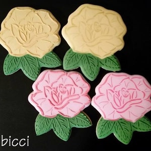 May include: Four sugar cookies decorated with icing in the shape of roses. Two cookies are yellow with green leaves and two cookies are pink with green leaves.