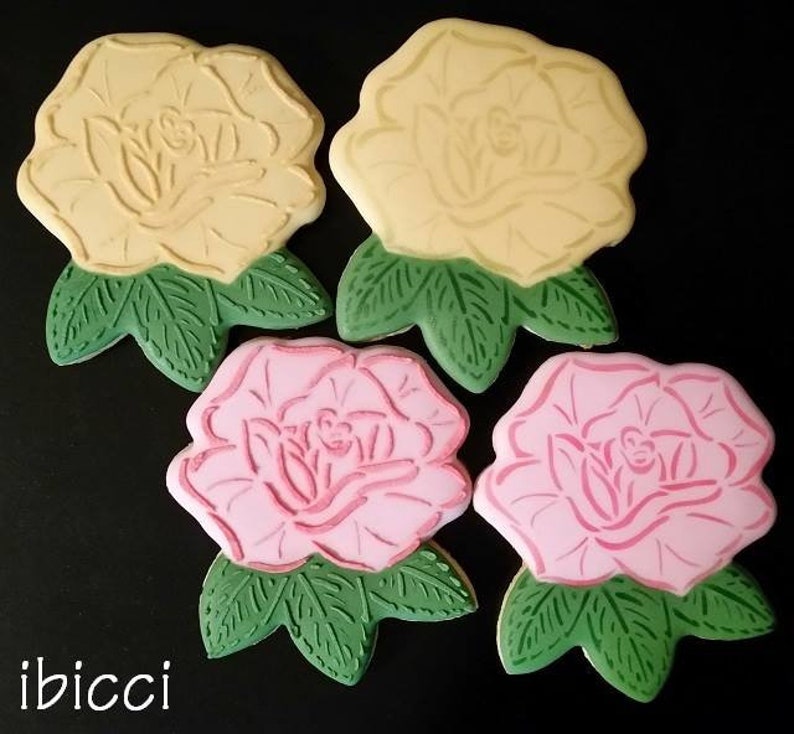 May include: Four sugar cookies decorated with icing in the shape of roses. Two cookies are yellow with green leaves and two cookies are pink with green leaves.