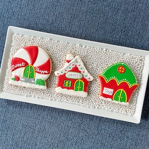 May include: Three Christmas-themed cookies on a white tray filled with white sprinkles. The cookies are shaped like a candy shop, a house with "Santa" on it, and a toy shop. The cookies are decorated with red, green, and white icing.