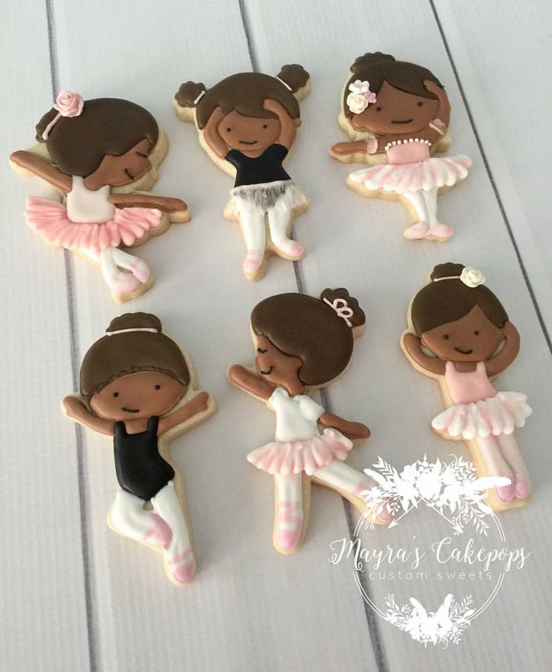 Op de afbeelding: Versierde suikerkoekjes in de vorm van ballerina's. Elk koekje toont een bruinharige ballerina in een roze tutu en balletschoenen. Sommige ballerina's dragen zwarte turnpakjes. De koekjes zijn gerangschikt op een wit oppervlak.