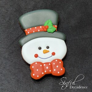 May include: A gingerbread cookie decorated with white icing and red and white polka dots. The cookie is shaped like a snowman with a black top hat and a green holly sprig.