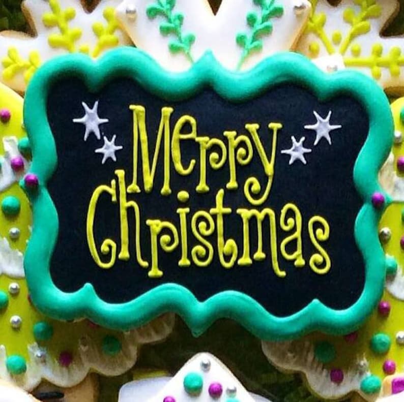 May include: A decorated cookie with a black background and the words "Merry Christmas" in yellow script. The cookie is framed with a green border and decorated with white star accents. Other cookies are visible in the background.
