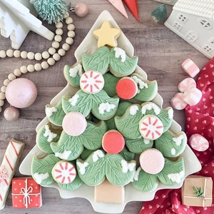 May include: A white ceramic Christmas tree-shaped plate with green and white frosted sugar cookies decorated with red and pink accents. The cookies are arranged in the shape of a Christmas tree.