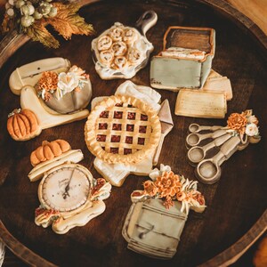 May include: An assortment of colourful and intricately decorated sugar cookies, including a pie, a scale, a measuring cup, a house, and a set of measuring spoons. The cookies are arranged on a wooden tray.