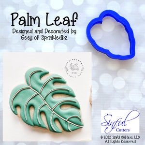 May include: A blue plastic cookie cutter in the shape of a palm leaf. The cutter is on a white background with a blurred image of a decorated cookie in the background.