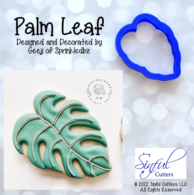 May include: A blue plastic cookie cutter in the shape of a palm leaf. The cutter is on a white background with a blurred image of a decorated cookie in the background.