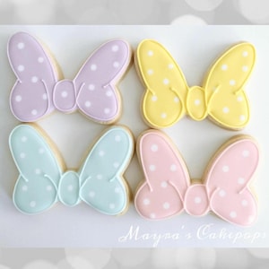 Peut inclure: Quatre biscuits en forme de papillon avec des pois blancs. Les biscuits sont de couleurs pastel : lavande, jaune, bleu et rose. Les biscuits ont un contour blanc et sont sur une surface blanche. Le texte "Mayna's Cakepops" est en bas.