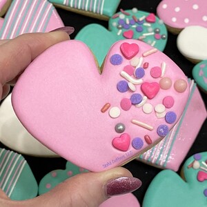 Puede incluir: Galletas en forma de corazón rosa decoradas con chispas blancas, rosas y moradas y pequeños corazones rosas. La galleta se sostiene en una mano y el texto "Simply Cutters, LLC" es visible en la esquina inferior derecha.