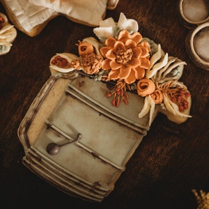 Op de afbeelding: Een versierde suikerkoek in de vorm van een vintage gieter, versierd met oranje, witte en groene bloemen glazuur. De koek heeft een lichtbruine rand en ligt op een donkere houten ondergrond. Andere koekjes zijn zichtbaar op de achtergrond.