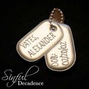 May include: Two silver dog tags engraved with "Yates, Alexander" and the date "02/20/02". They are linked by a ball chain and a brown leather-like piece. The background is black. The image also includes the text "Sinful Decadence".