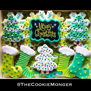May include: A box of decorated Christmas cookies. The cookies include Christmas trees, snowflakes, and stockings, all in green, white, and yellow icing. A central cookie reads "Merry Christmas" in yellow script on a black background.