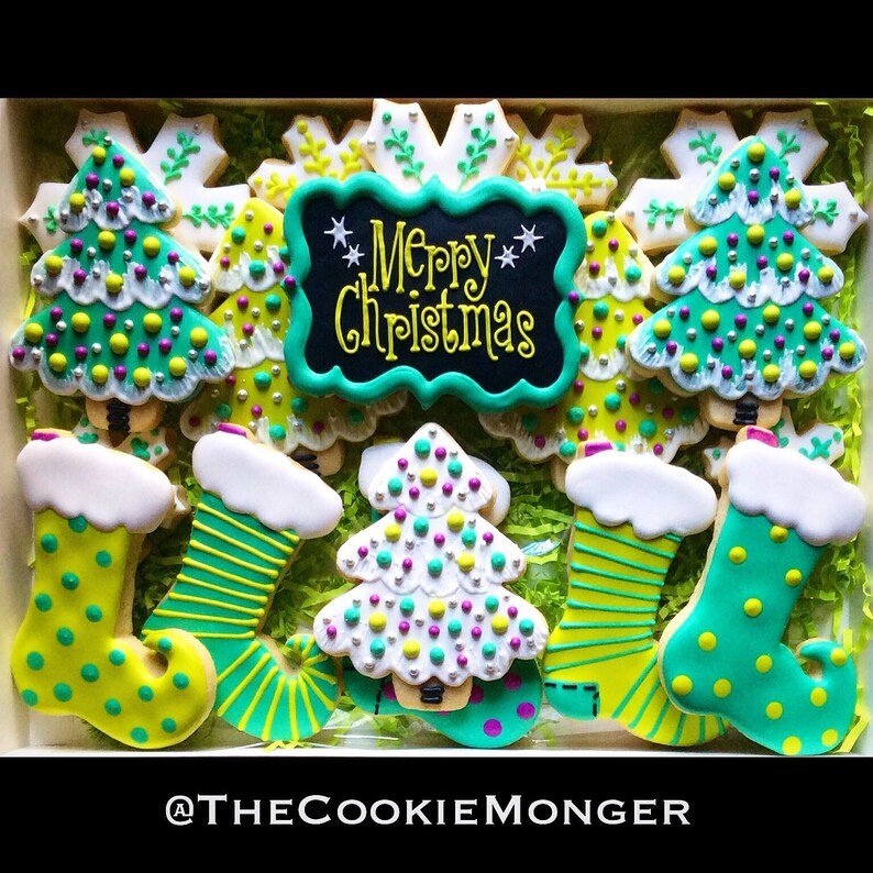 May include: A box of decorated Christmas cookies. The cookies include Christmas trees, snowflakes, and stockings, all in green, white, and yellow icing. A central cookie reads "Merry Christmas" in yellow script on a black background.