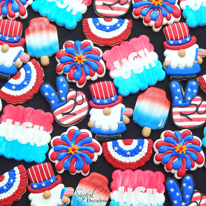 May include: A collection of decorated sugar cookies featuring patriotic themes, including fireworks, gnomes, peace signs, and popsicles. The cookies are decorated in red, white, and blue with stars and stripes.