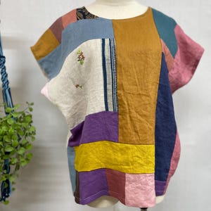 May include: A short-sleeved top constructed from various rectangular fabric patches in mustard yellow, navy blue, light blue, pink, purple, and beige. The beige section features small embroidered floral details and a vertical blue striped panel.