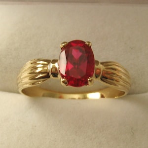 May include: A gold ring featuring a large, oval-shaped, red gemstone. The ring band has a textured design.