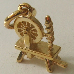 May include: A gold-colored spinning wheel charm with a detailed design. The wheel is mounted on a small wooden base with a spindle.