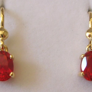 May include: A pair of gold earrings with red oval-shaped gemstones. The earrings have a simple design with a small gold ball at the top of the hook.
