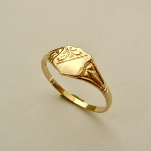 May include: A gold signet ring with a decorative design on the top. The ring is made of solid gold and has a smooth finish.