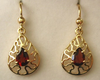 May include: A pair of gold-colored drop earrings. Each earring features a teardrop-shaped, openwork design with a vibrant, red gemstone at the center. The earrings have a hook closure and a small, round accent at the top.