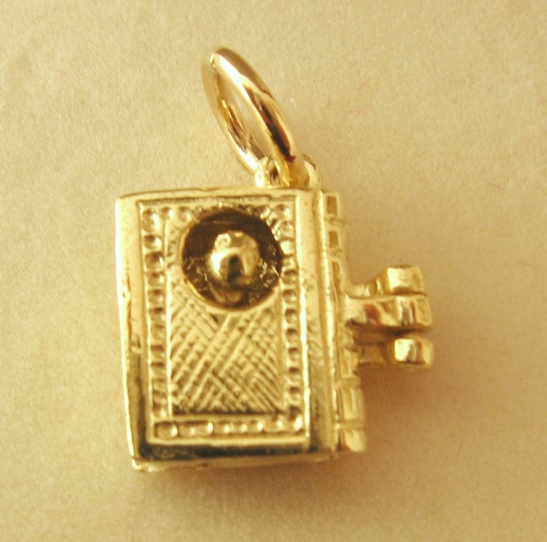 May include: A gold-toned charm in the shape of a closed book with a small round knob on the front. The charm has a hinged clasp on the side.