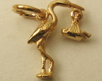 May include: Gold-toned charm of a stork carrying a baby in a swaddle. The stork is standing on a base with a loop for attaching to a necklace or bracelet.