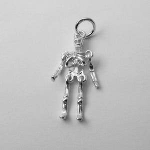 May include: A silver-colored metal skeleton charm featuring articulated joints at the shoulders and hips. The figure has a detailed rib cage, a skull head, and long limbs, with a circular jump ring attached to the top of the skull for hanging.