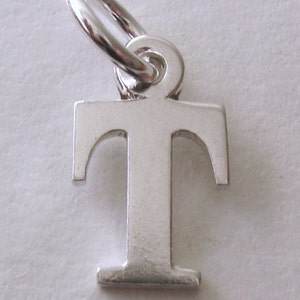 May include: Silver charm in the shape of the letter T. The charm has a loop at the top for attaching to a bracelet or necklace.