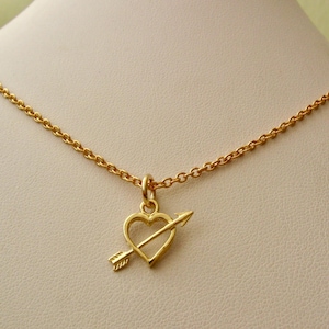 May include: A gold necklace with a heart pendant pierced by an arrow. The heart is open and the arrow is pointing to the right.
