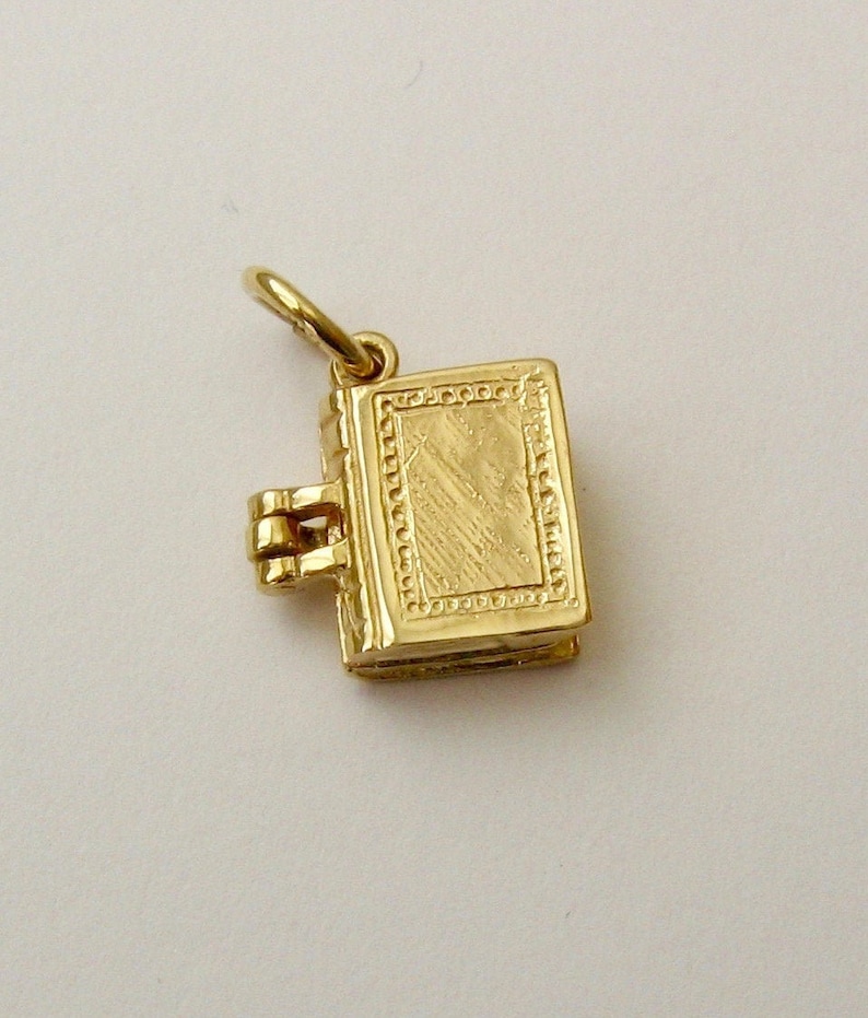 May include: A gold-colored book charm with a hinged clasp. The charm is detailed with a textured surface and a decorative border.
