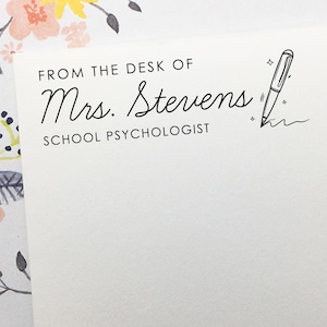 May include: A white notepad with the text "FROM THE DESK OF Mrs. Stevens SCHOOL PSYCHOLOGIST" in black script. A pen illustration is next to the text. The notepad is on a white surface with floral accents.