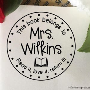 Puede incluir: Sello redondo blanco y negro con el texto "This book belongs to Mrs. Wilkins Read it, love it, return it!" y un icono de libro abierto.