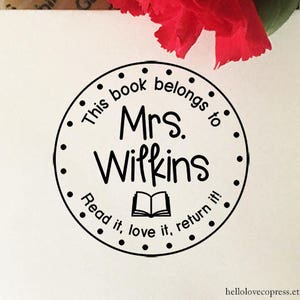Puede incluir: Sello redondo en blanco y negro con el texto "This book belongs to Mrs. Wilkins Read it, love it, return it!" y una imagen de un libro abierto.