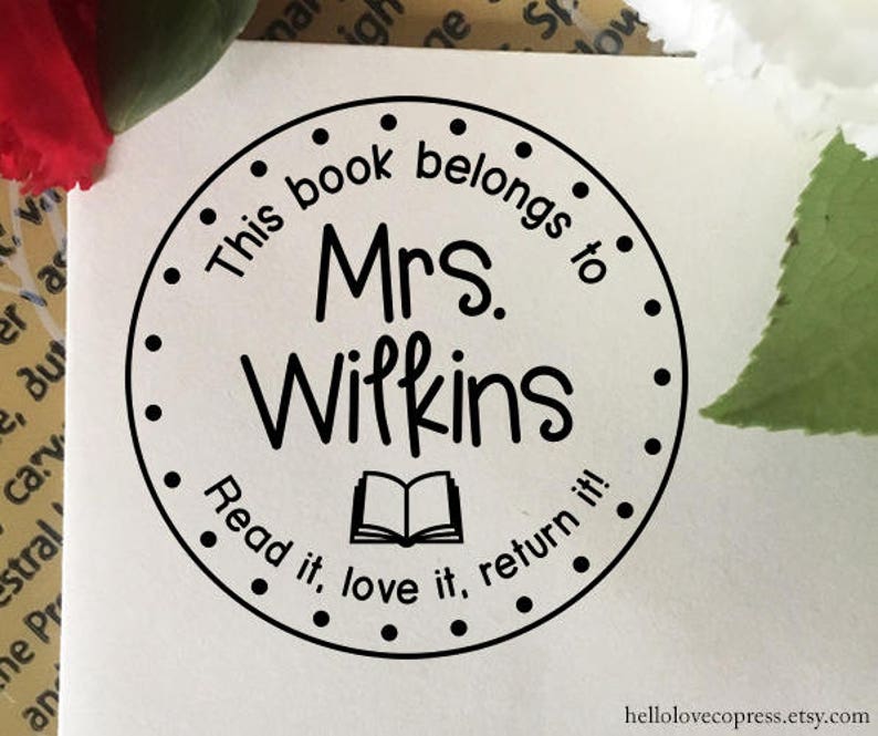 Puede incluir: Sello redondo blanco y negro con el texto "This book belongs to Mrs. Wilkins Read it, love it, return it!" y un icono de libro abierto.