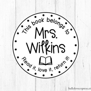 Puede incluir: Sello circular blanco y negro con el texto "This book belongs to Mrs. Wilkins Read it, love it, return it!" y una imagen de un libro abierto.