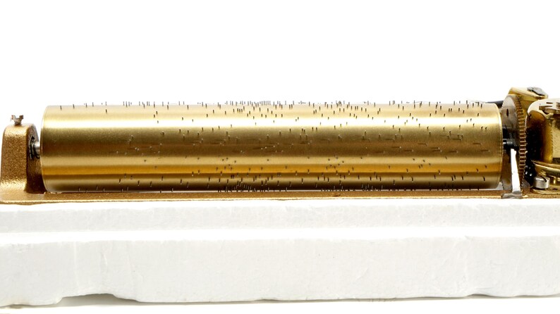 May include: A vintage music box mechanism with a gold-colored cylinder covered in small pins. The mechanism is mounted on a white base, and the gears and other components are also gold-toned. This antique music box is a collectible item.