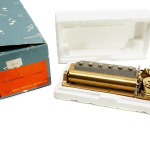 May include: A vintage Swiss musical movement, featuring a gold-toned metal mechanism with a series of pins and a cylinder. The movement is displayed in a white foam insert, next to its original blue box with text and a red label.