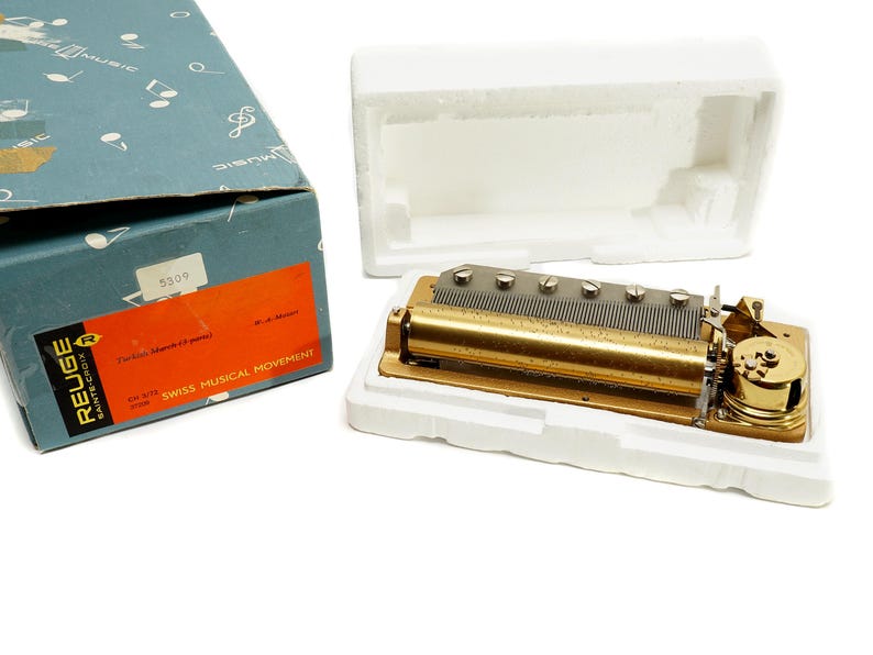 May include: A vintage Swiss musical movement, featuring a gold-toned metal mechanism with a series of pins and a cylinder. The movement is displayed in a white foam insert, next to its original blue box with text and a red label.
