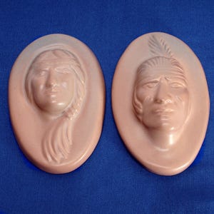 May include: Two oval-shaped, pink-toned wall plaques. Each plaque features a sculpted face of a Native American person. One has a woman's face with long hair, the other a man's face with a feather in his hair.