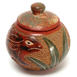May include: A round, lidded ceramic jar featuring a textured tan surface with a carved and painted red lizard and green leaves. The lid has a matching red and green mottled finish with a central knob handle, creating a decorative container.