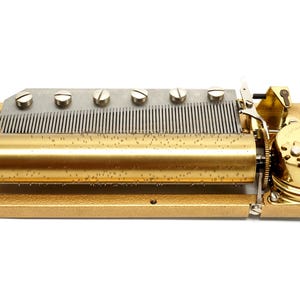 May include: A vintage music box mechanism with a gold-colored frame and cylinder. The mechanism features a series of metal tines, screws, and gears. The cylinder is covered in small pins. The music box is designed to play a melody.