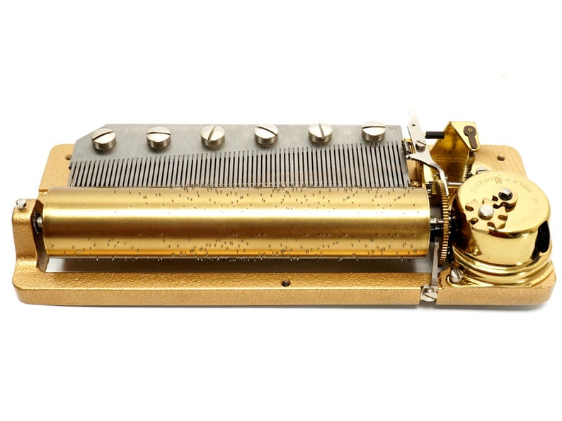 May include: A vintage music box mechanism with a gold-colored frame and cylinder. The mechanism features a series of metal tines, screws, and gears. The cylinder is covered in small pins. The music box is designed to play a melody.