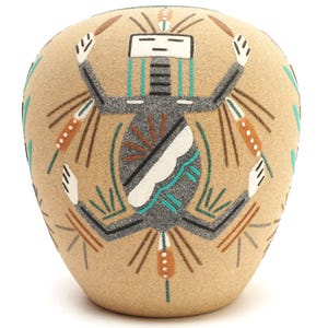 May include: A tan ceramic vase featuring a textured surface with a central stylized figure in black, white, and turquoise. The figure has raised arms and is surrounded by geometric lines and patterns in brown, white, and turquoise accents.