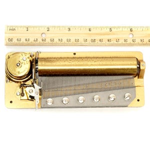 May include: A vintage music box mechanism with a brass finish. The mechanism includes a cylinder, comb, and a series of screws. A wooden ruler is placed above the mechanism to show the size. The mechanism is approximately 6 inches long.