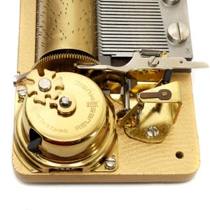 May include: Close-up of a vintage music box mechanism. The brass components include a cylinder with pins, gears, and a bell-shaped housing. The text "REUGE MUSIC SWITZERLAND" is visible on the housing. The mechanism is mounted on a wooden base.