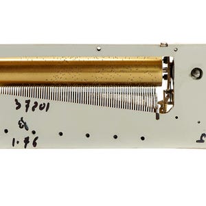 May include: A vintage music box mechanism with a brass-colored cylinder and a series of metal tines. The mechanism is mounted on a rectangular, light gray metal plate. The text "37201" and "1.46" are handwritten on the plate.