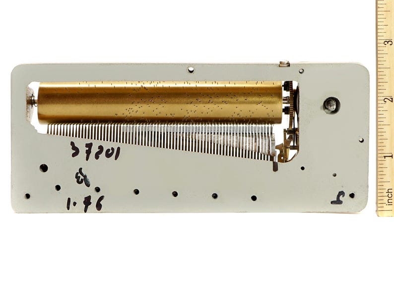 May include: A vintage music box mechanism with a brass-colored cylinder and a series of metal tines. The mechanism is mounted on a rectangular, light gray metal plate. The text "37201" and "1.46" are handwritten on the plate.
