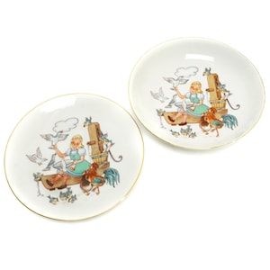 May include: Two white porcelain plates with a gold rim. Each plate features a colorful illustration of a girl feeding birds and a rooster. The plates are perfect for serving snacks or desserts.